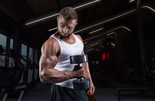 Muscular man in a white t-shirt works out in the gym with dumbbells. Biceps pumping. Fitness and bodybuilding concept.