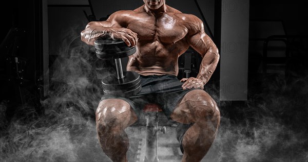 Muscular man sits on a bench in smoke with dumbbells. Bodybuilding and powerlifting concept.