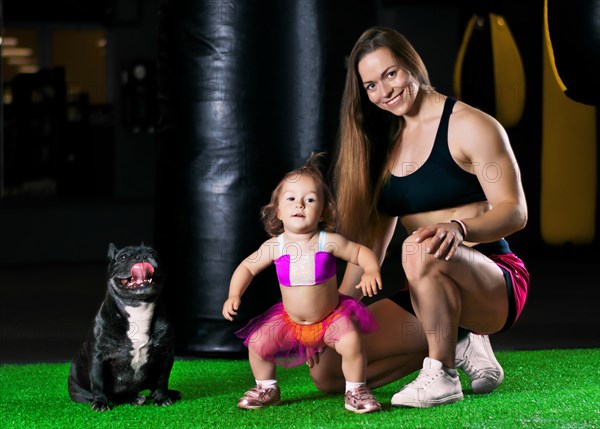 Charming sports mom trains in the gym with her little daughter and a French bulldog.