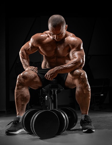 Muscular man sits on a bench near huge dumbbells. Bodybuilding and powerlifting concept.