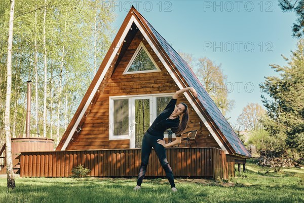 Sporty woman does side body bent on lawn of a log cabin in the morning