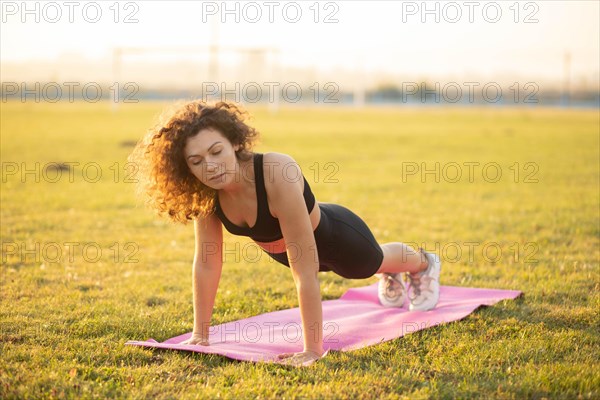 Young curly athletic girl in sportswear performs a plank exercise on a yoga mat outdoors on the grass during sunset