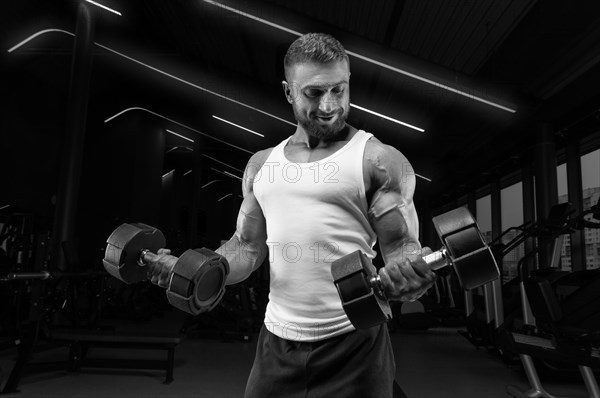 Muscular man in a white t-shirt works out in the gym with dumbbells. Biceps pumping. Fitness and bodybuilding concept.