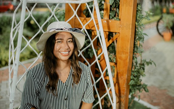 Portrait of a smiling girl in a hat sitting on a white swing in a garden. Lifestyle of girl in hat sitting on a swing looking at camera