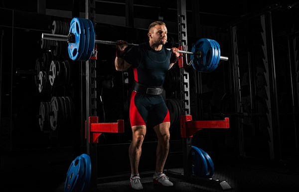 The weightlifter stands with a very heavy barbell on his shoulders. He is at the top and is preparing to sit down