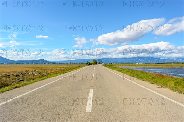 A straight road leading towards mountains under a bright blue sky with clouds