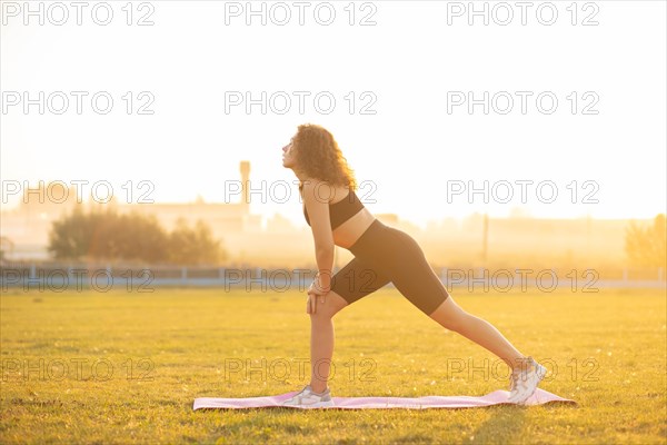 Young curly athletic girl in sportswear doing leg exercises outdoors during sunset