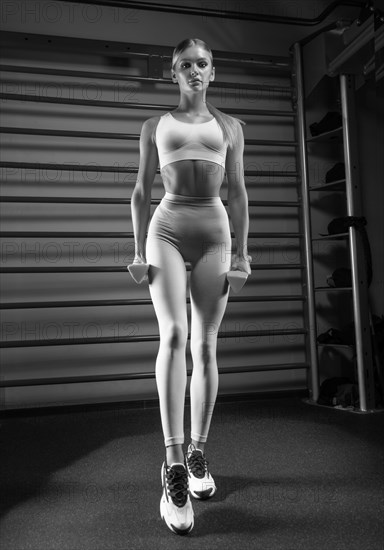 Beautiful tall blonde posing in the gym with dumbbells in her hands against the background of the wall bar. The concept of sports