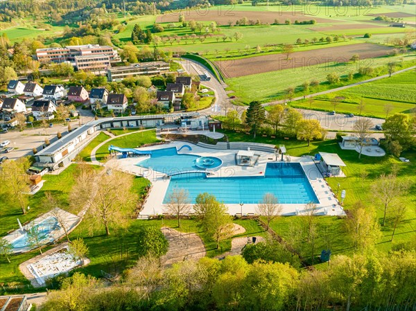 Wide-angle drone view of an outdoor swimming pool surrounded by green landscape in the sunshine