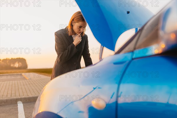 A young beautiful stylish girl driver in a jacket is trying to identify the cause of a car breakdown under the hood