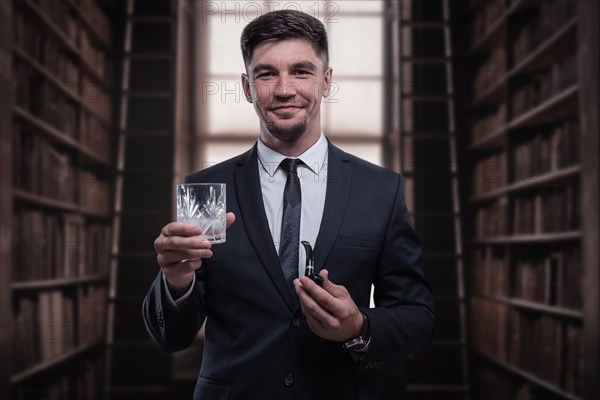 Portrait of an elegant man in a suit with a smoking pipe and a glass in the library. Business concept.