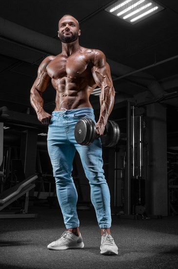 Pumped-up man posing in the gym in jeans with a dumbbell in his hand. Sports