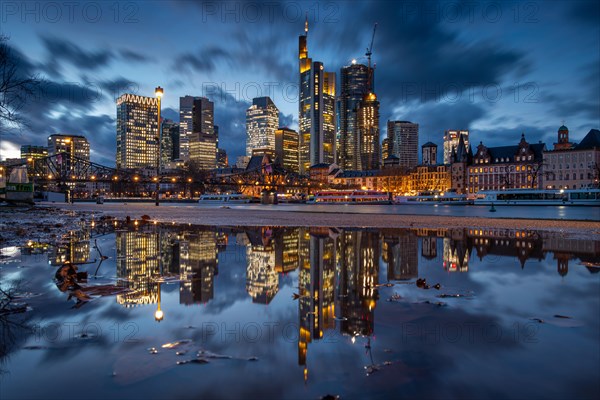 The glowing Frankfurt bank skyline is reflected in a puddle on the banks of the Main in the evening