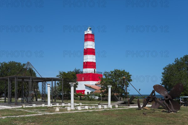Red and white lighthouse under a blue sky next to green areas and a historic anchor