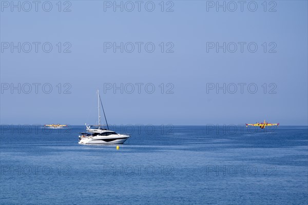 Two fire-fighting aircraft refuelling water at the sea with sailing yacht in the foreground