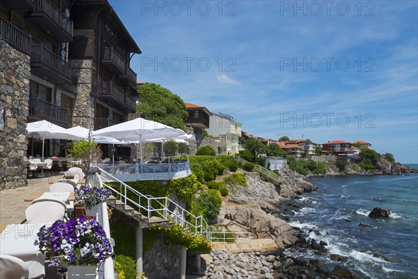 Stone building next to a clear blue sea with a terrace and white chairs