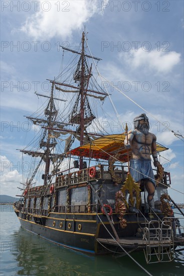 A large sailing ship with many masts and a pirate statue on board is anchored in the sea