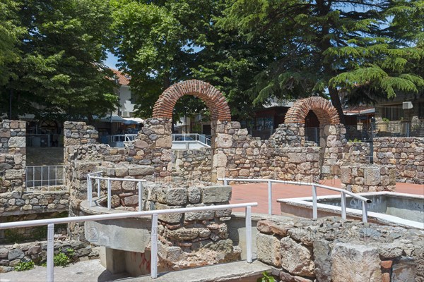 Ruins of an ancient structure with brick arches in an urban setting under a blue sky