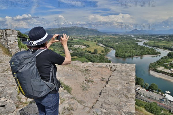 Woman in a sun hat takes a photo with her mobile phone of the view from the Illyrian fortress Rozafa to the river Drin