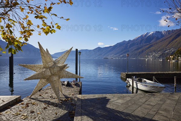 Backlit photograph of the large Herrenhuth star as a Christmas decoration at the harbour entrance to Lake Maggiore with lanterns and motorboat in Ascona