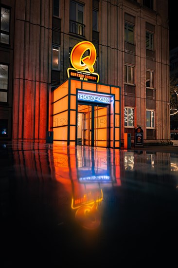 A theatre box office at night with a glowing neon sign and its reflection in the floor