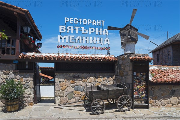 Traditional restaurant building with windmill and wooden wagon under a clear blue sky