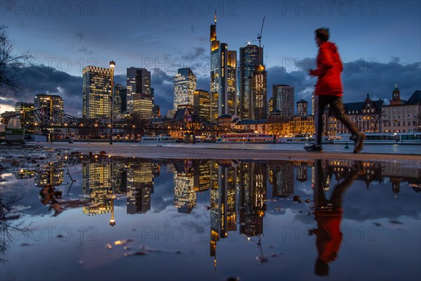 The glowing Frankfurt bank skyline is reflected in a puddle on the banks of the Main as a jogger runs past