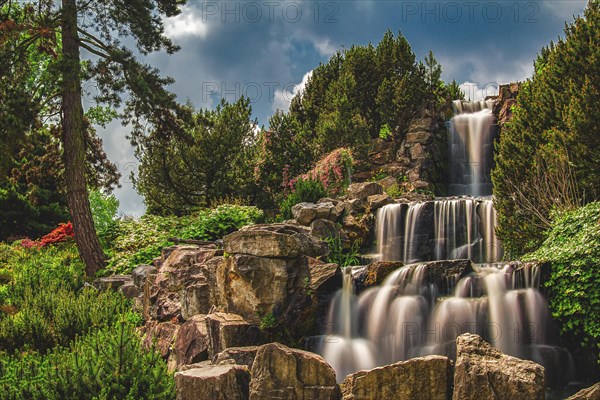 A picturesque waterfall flows over rocky steps surrounded by lush vegetation and dramatic skies, Gruga, Essen, North Rhine-Westphalia, Germany, Europe