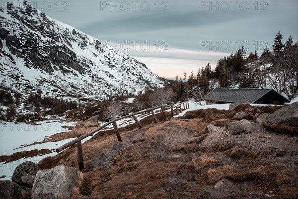 A serene winter landscape with a cabin, snow-covered mountain, and wooden fence at dusk. Poland, Karkonosze