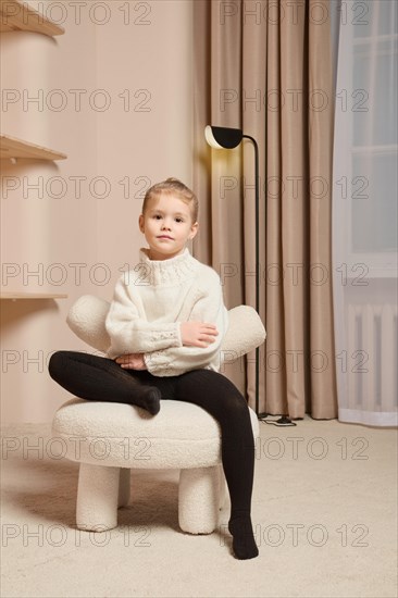 A little girl in warm sweater sits on a soft chair with one leg crossed and her arms crossed over her chest