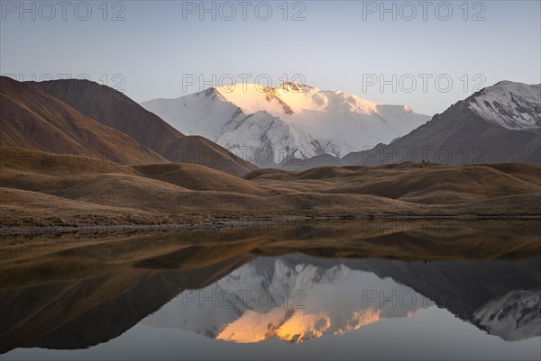 White glaciated and snowy mountain peak Pik Lenin at sunset, mountains reflected in a lake between golden hills, Trans Alay Mountains, Pamir Mountains, Osh Province, Kyrgyzstan, Asia