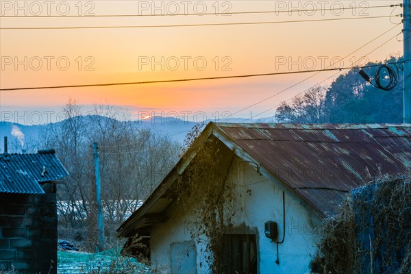 Old stucco house with rusted tin roof with sun setting behind mountains bouncing off the atmosphere and smoke from chimney in background