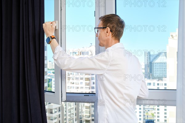 Middle aged man opens window in his apartment of high-rise building