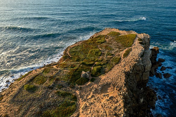 Drone view of rocky headland and blue sea waves at sunset. Ocean tide beat and splash in summer sunset haze, little foliage and rocky cliffs, Peniche, Portugal, Europe
