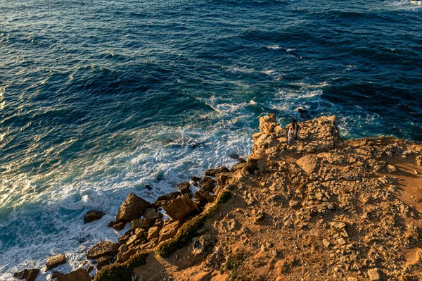 Aerial landscape of rocky shores and blue ocean waves. Drone shot of sea blue waves beat and splash in summer sunset haze, little foliage and rocky cliffs, establishing or static shot, Peniche, Portugal, Europe