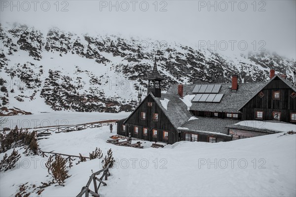 Wooden cabin covered in snow with mountains in the background on a cloudy day. Shelter in the mountains