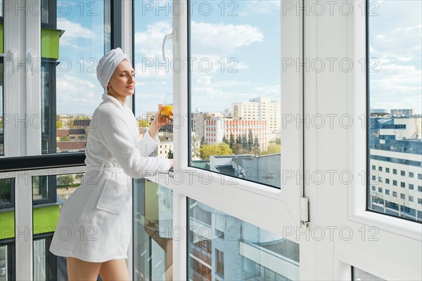 Young woman in a bathrobe stands on balcony of her new apartment and squint eyes