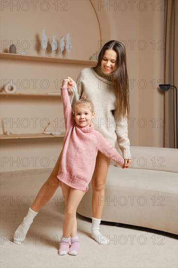 A joyful mother and her young daughter dancing together in a cozy living room. The daughter wears a pink sweater, while the mother is in a grey sweater