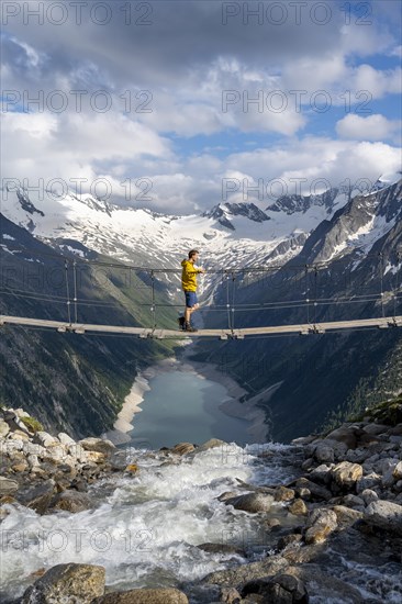 Mountaineers on a suspension bridge over a mountain stream Alelebach, picturesque mountain landscape near the Olpererhuette, view of turquoise-blue lake Schlegeisspeicher, glaciated rocky mountain peaks Hoher Weisszint and Hochfeiler with glacier Schlegeiskees, Berliner Hoehenweg, Zillertal Alps, Tyrol, Austria, Europe