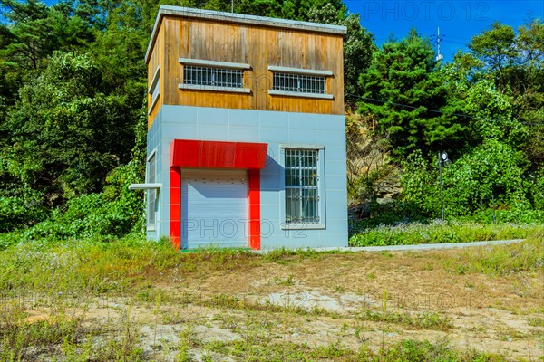 Square house with red awning over garage door in wooded area surrounded by green trees and flowers