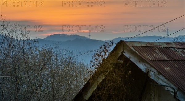 Old house with rusted tin roof with sun setting behind mountains bouncing off the atmosphere and smoke from chimney in background