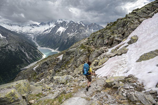 Mountaineer on hiking trail with snow, view of Schlegeisspeicher, glaciated rocky mountain peaks Hoher Weisszint and Hochfeiler with glacier Schlegeiskees, Berliner Hoehenweg, Zillertal Alps, Tyrol, Austria, Europe