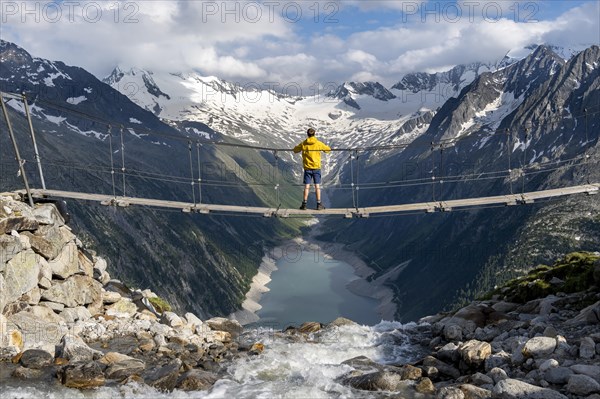 Mountaineers on a suspension bridge over a mountain stream Alelebach, picturesque mountain landscape near the Olpererhuette, view of turquoise-blue lake Schlegeisspeicher, glaciated rocky mountain peaks Hoher Weisszint and Hochfeiler with glacier Schlegeiskees, Berliner Hoehenweg, Zillertal Alps, Tyrol, Austria, Europe