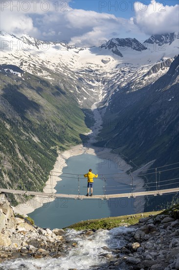 Mountaineers on a suspension bridge over a mountain stream Alelebach, picturesque mountain landscape near the Olpererhuette, view of turquoise-blue lake Schlegeisspeicher, glaciated rocky mountain peaks Grosser Moeseler, Hoher Weisszint and Hochfeilermit glacier Schlegeiskees, Berliner Hoehenweg, Zillertal Alps, Tyrol, Austria, Europe