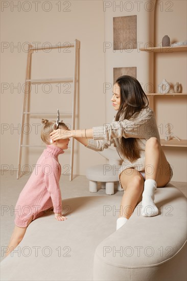 A mother and daughter are playing together in a modern living room. Daughter asks her mother to put on her a hair hoop
