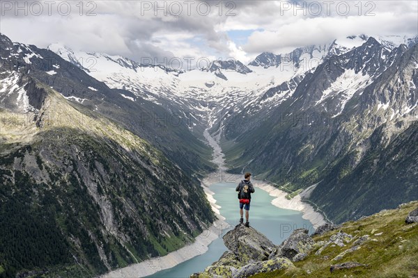 Mountaineer on a rock in front of a mountain panorama, view of Schlegeisspeicher, glaciated rocky mountain peaks Hoher Weisszint and Hochfeiler with glacier Schlegeiskees, Berliner Hoehenweg, Zillertal Alps, Tyrol, Austria, Europe