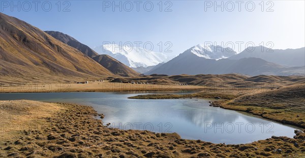 White glaciated and snow-covered mountain peak Pik Lenin at sunset, mountains reflected in Lake Tulpar-Kul, yurts between golden hills, Trans Alay Mountains, Pamir Mountains, Osh Province, Kyrgyzstan, Asia