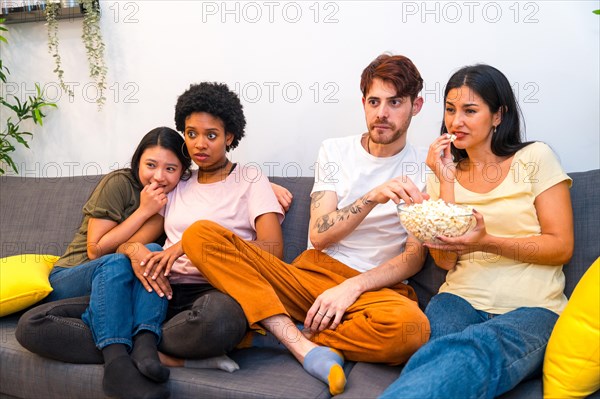 Friends eating pop corn while watching a thriller film together sitting on the sofa at home