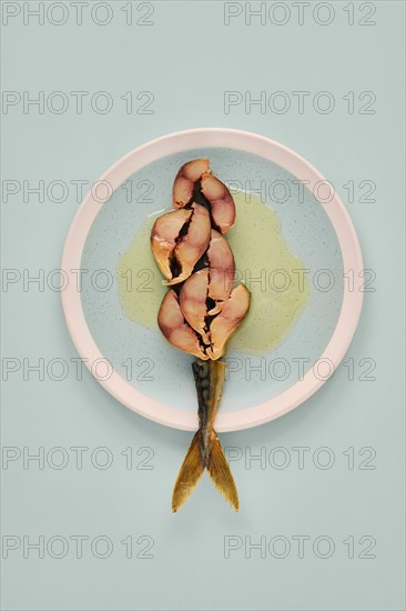Sliced mackerel on a plate revealing its pinkish inner flesh against the darker outer skin. The plate contrasts with both the light blue background and the colors of the fish and sauce