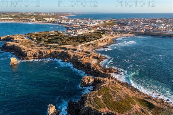 Drone view of rocky peninsula and town of Peniche, Portugal. Summer sunset haze, little foliage and rocky cliffs, peninsula and rocks, fishing town, horizon, ocean waves break on rocky shores